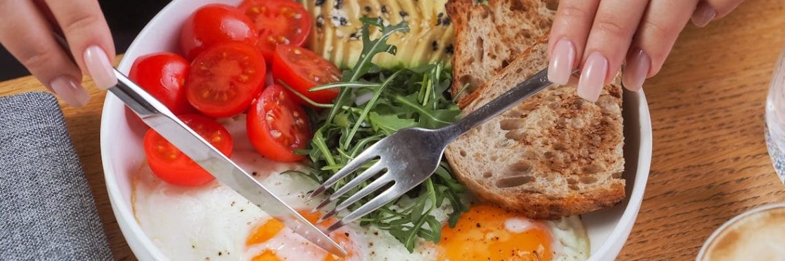 Close-up of a breakfast bowl with fried eggs, avocado, tomatoes, and bread, served with cappuccino.